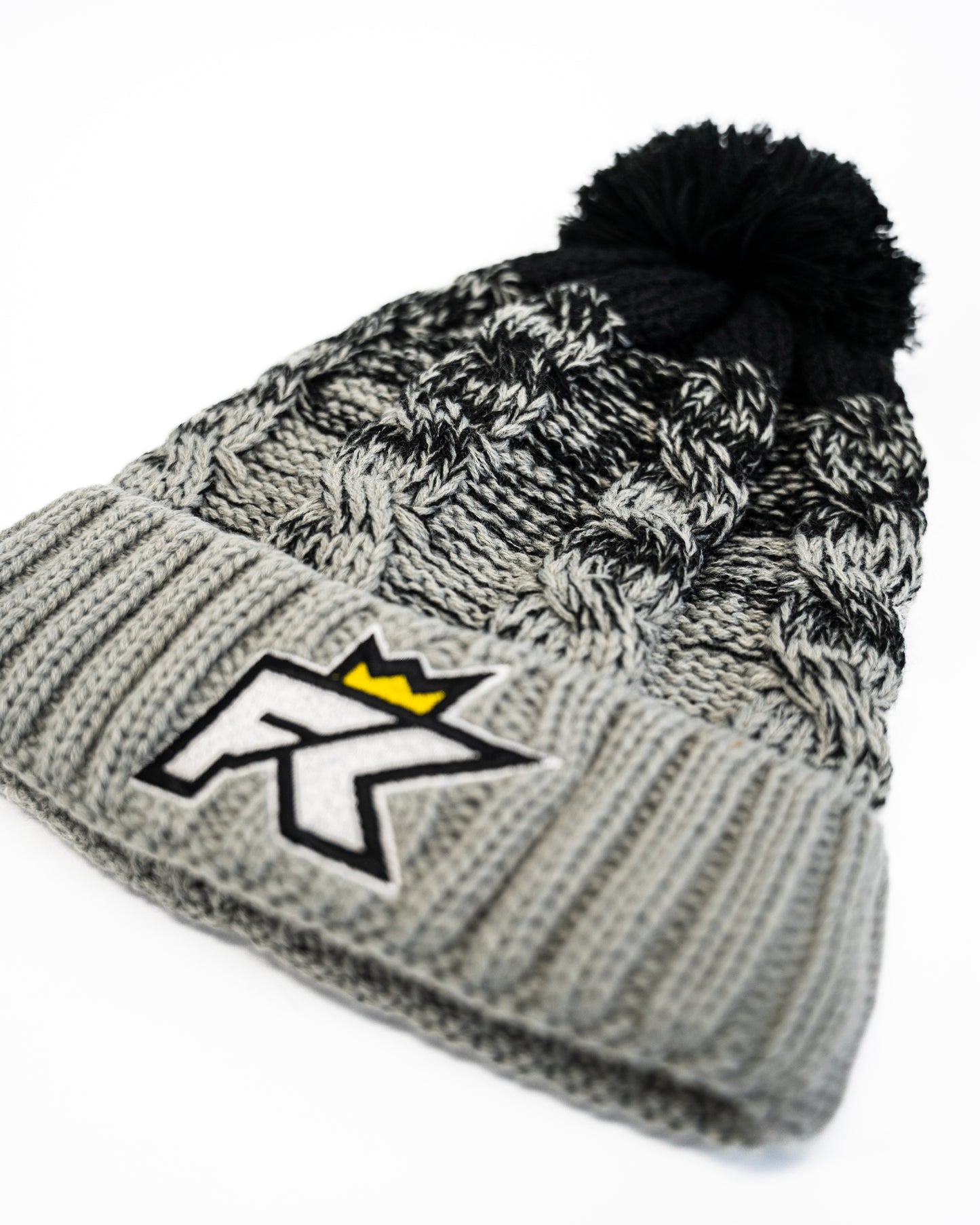 The FibreKing Knitted Bobble Hat features a heavyweight multi-yarn gray and black knit, a pom-pom, and an embroidered FK logo with a yellow crown on the front.