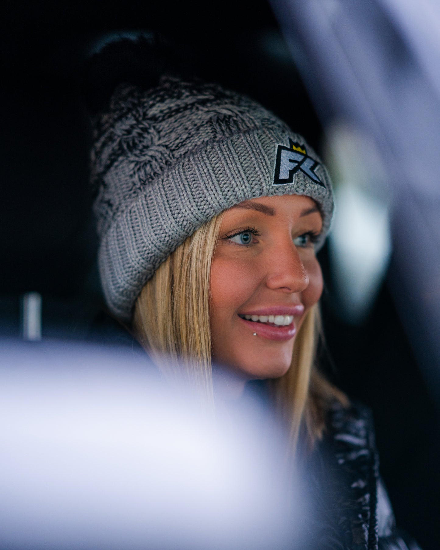 A woman in a gray FibreKing Knitted Bobble Hat and black jacket smiles indoors, with part of the view obscured by a blurred foreground.