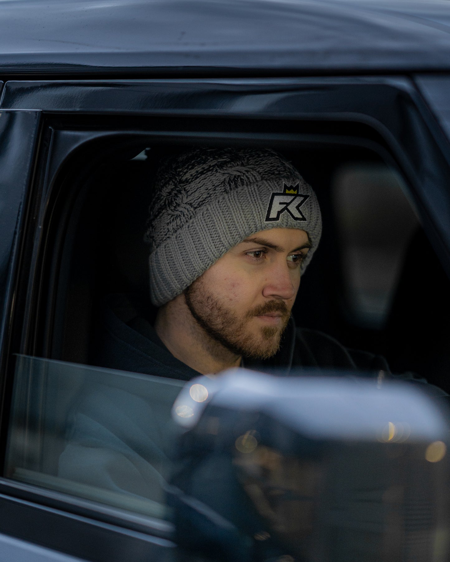 A bearded man wearing a grey FibreKing Knitted Bobble Hat sits in a vehicles driver seat, looking ahead with a neutral expression.