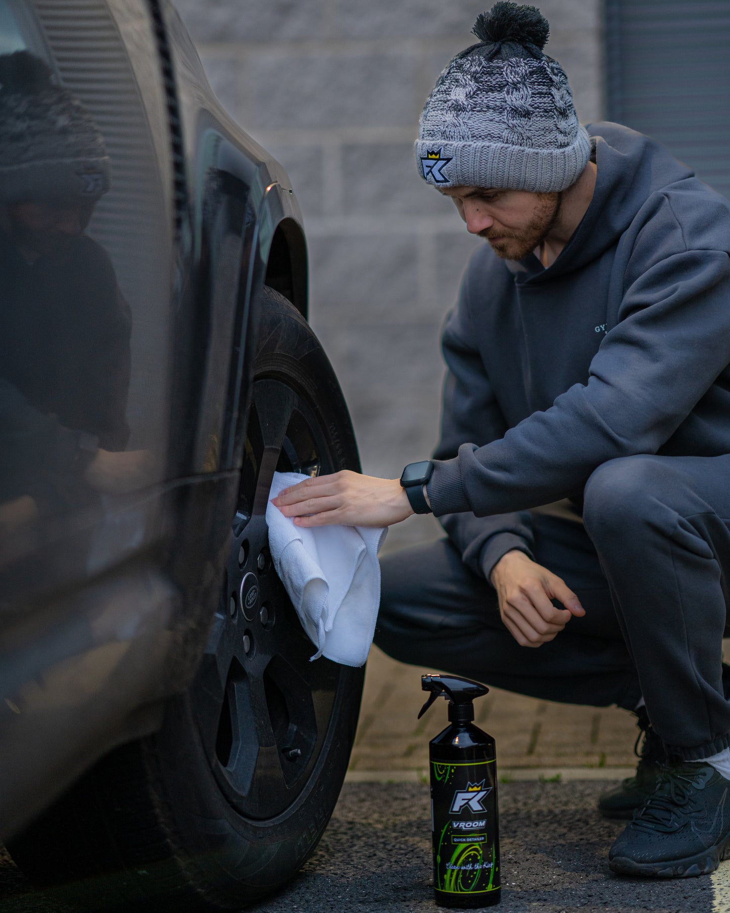 A person in a gray tracksuit and a FibreKing Fibreking Knitted Bobble Hat with polyester shearling lining crouches beside a car, cleaning the wheel with a white cloth while a bottle of wheel cleaner sits on the ground nearby.