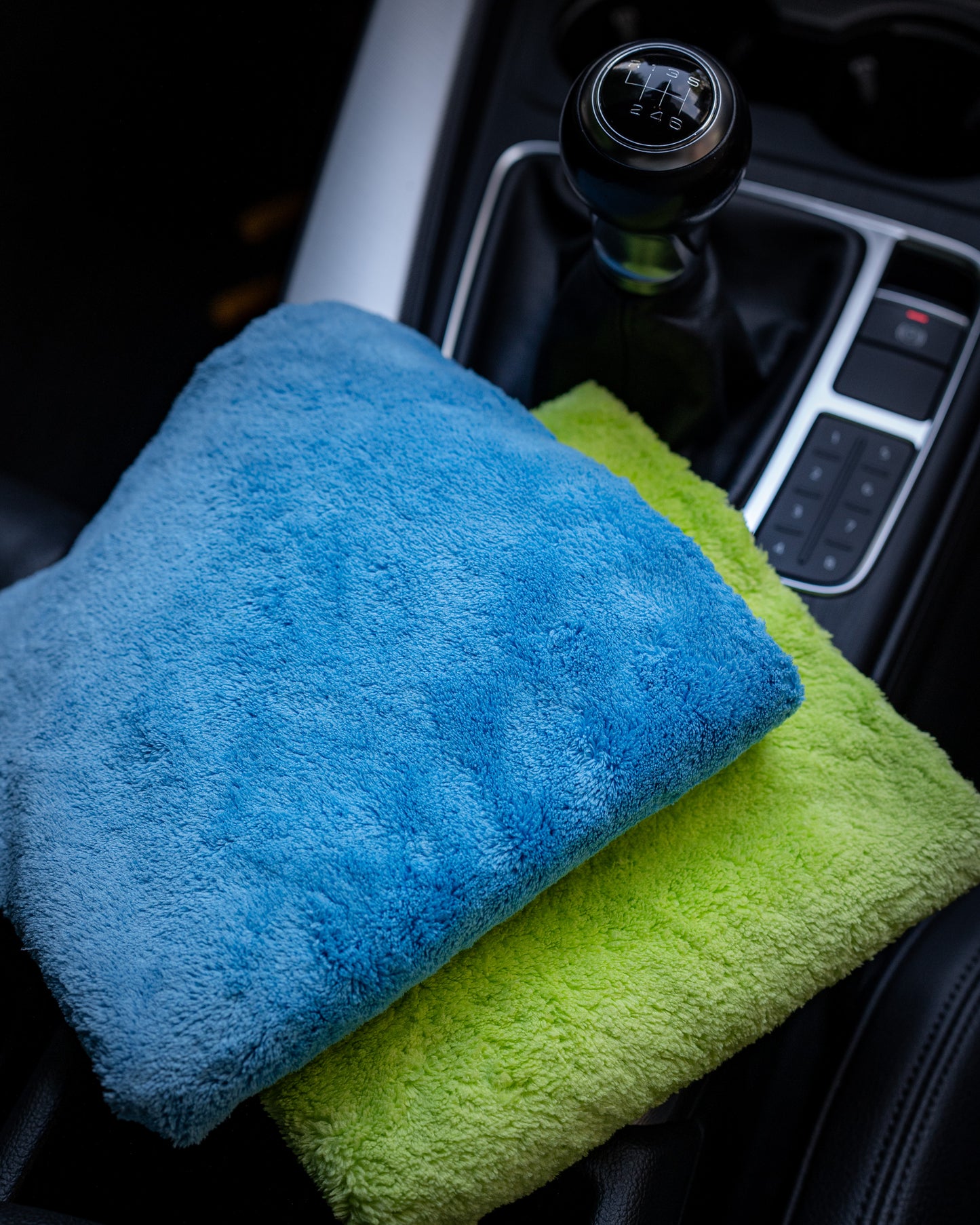 Two Ultimate detailer cloths by FibreKing—one blue, one yellow-green—made from coral fleece microfibre, are placed on a car’s center console near the gear shift.