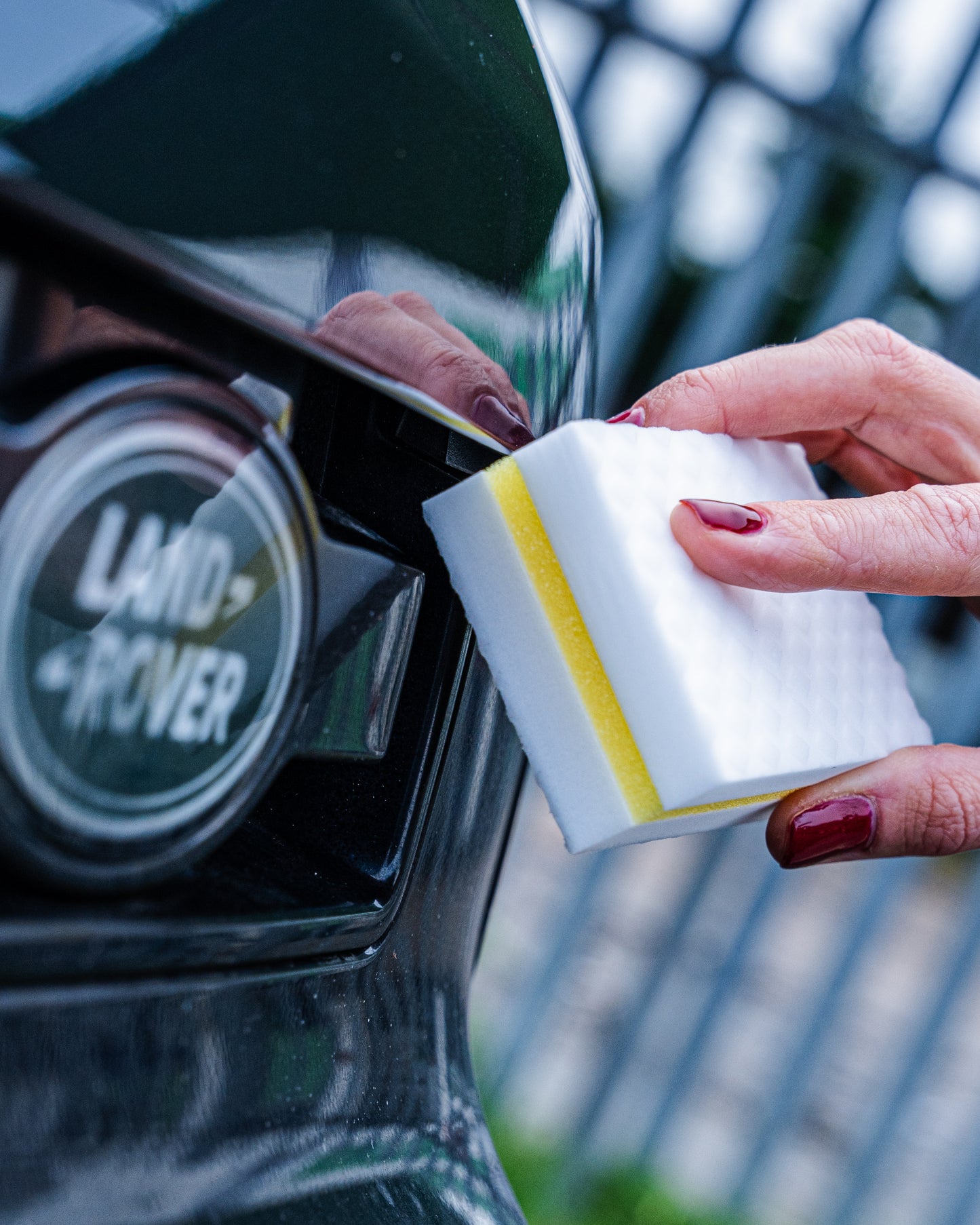 A person with red nail polish uses a FibreKing Magic Eraser melamine foam sponge (10 per pack) to clean the rear of a black Land Rover.