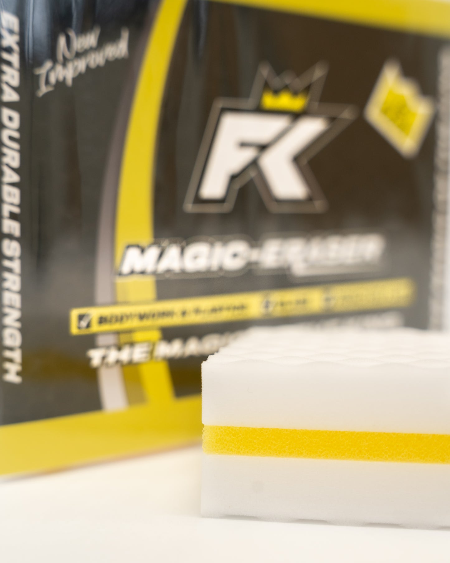A white melamine foam cleaning sponge with a yellow stripe sits before a black and yellow FibreKing Magic Eraser box (10 per pack)—perfect for tackling tough stains or motorsport cleaning.