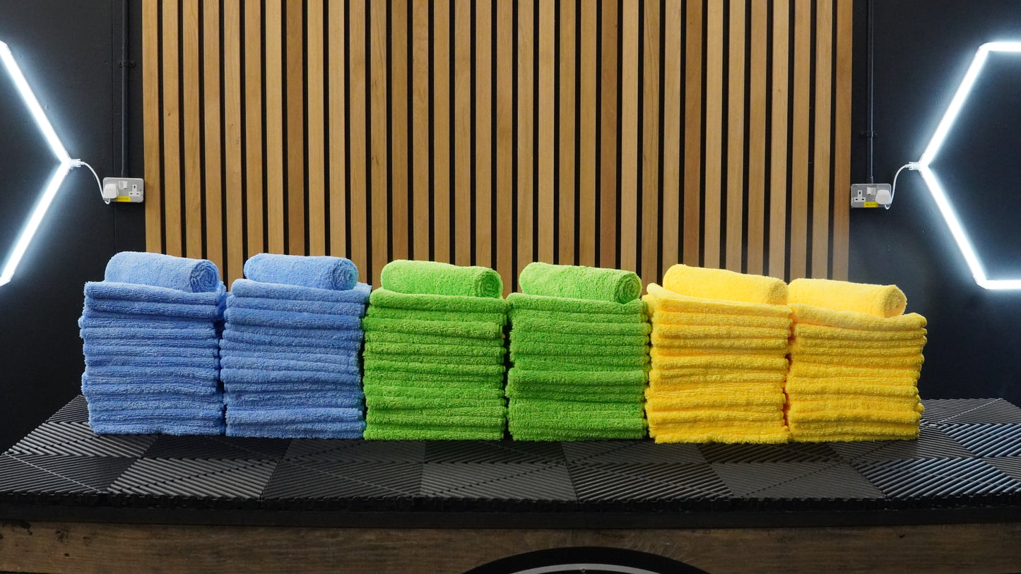 Stacks of blue, green, and yellow FibreKing Silver Label Microfibre Bundle cloths are neatly arranged on a black surface in front of wooden slats and geometric light fixtures.