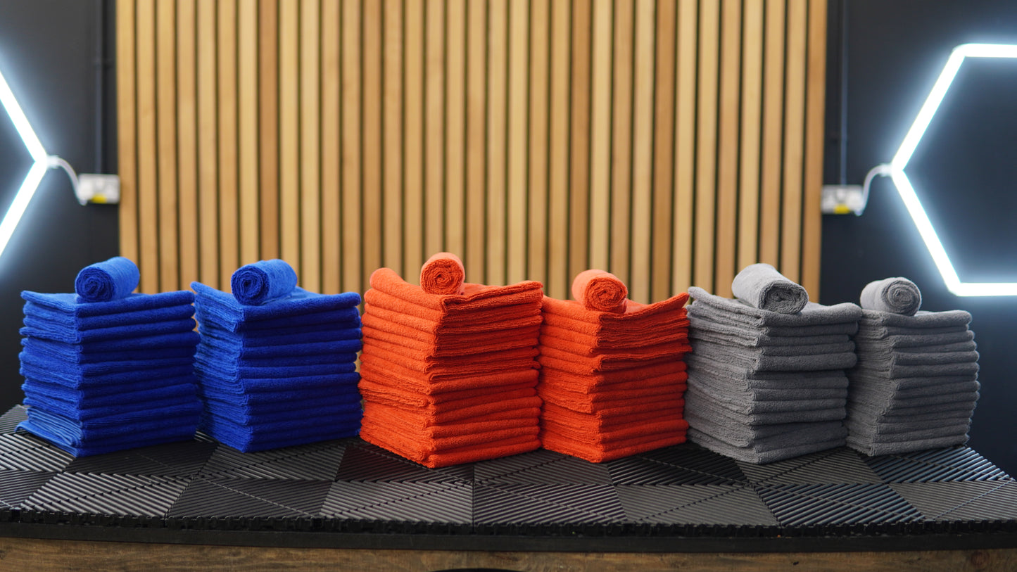 Stacks of neatly folded blue, orange, and gray FibreKing Gold Label Microfibre Bundle cloths, each topped with a rolled towel, are arranged on a black surface with a wooden slat background and geometric lighting.
