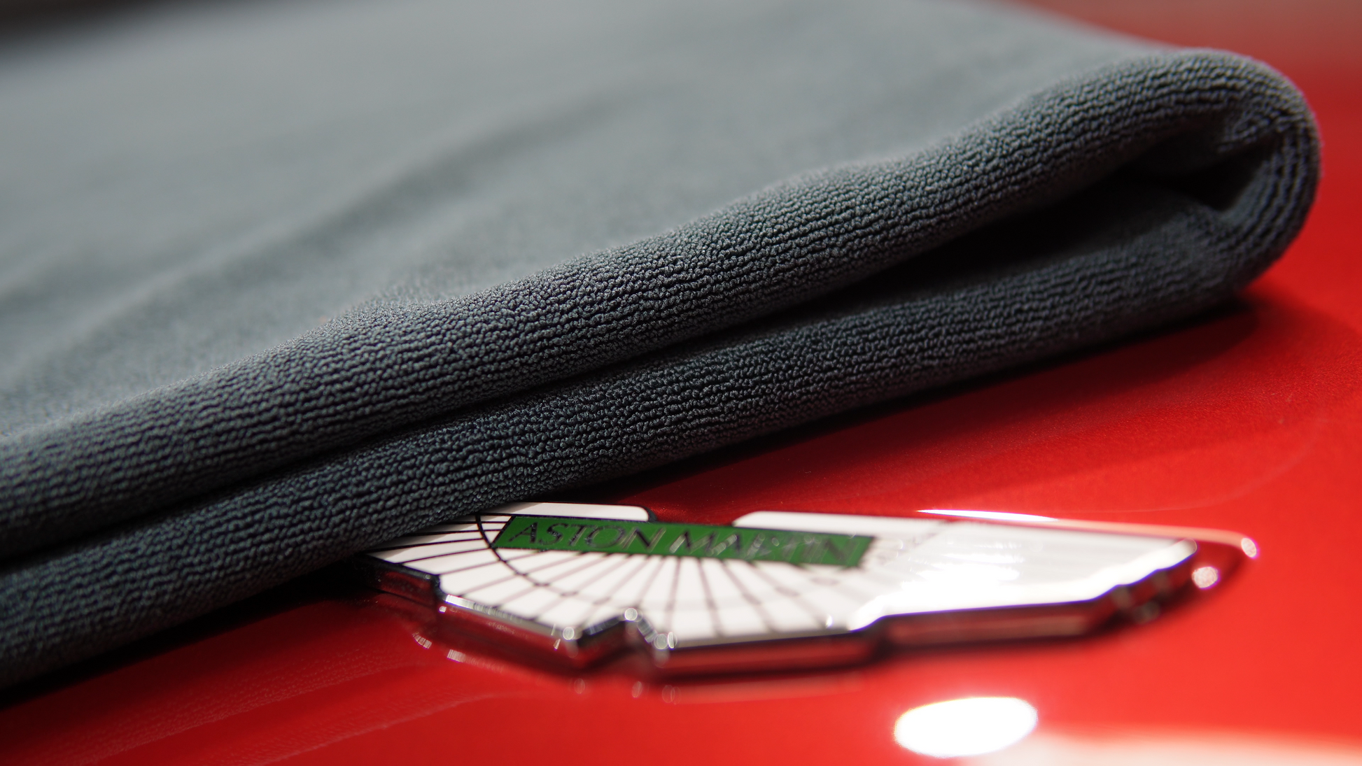 A FibreKing Gold Label 900 GSM Korean Drying Towel in gray partially covers the silver and green Aston Martin emblem on a shiny red car, showcasing premium car care.