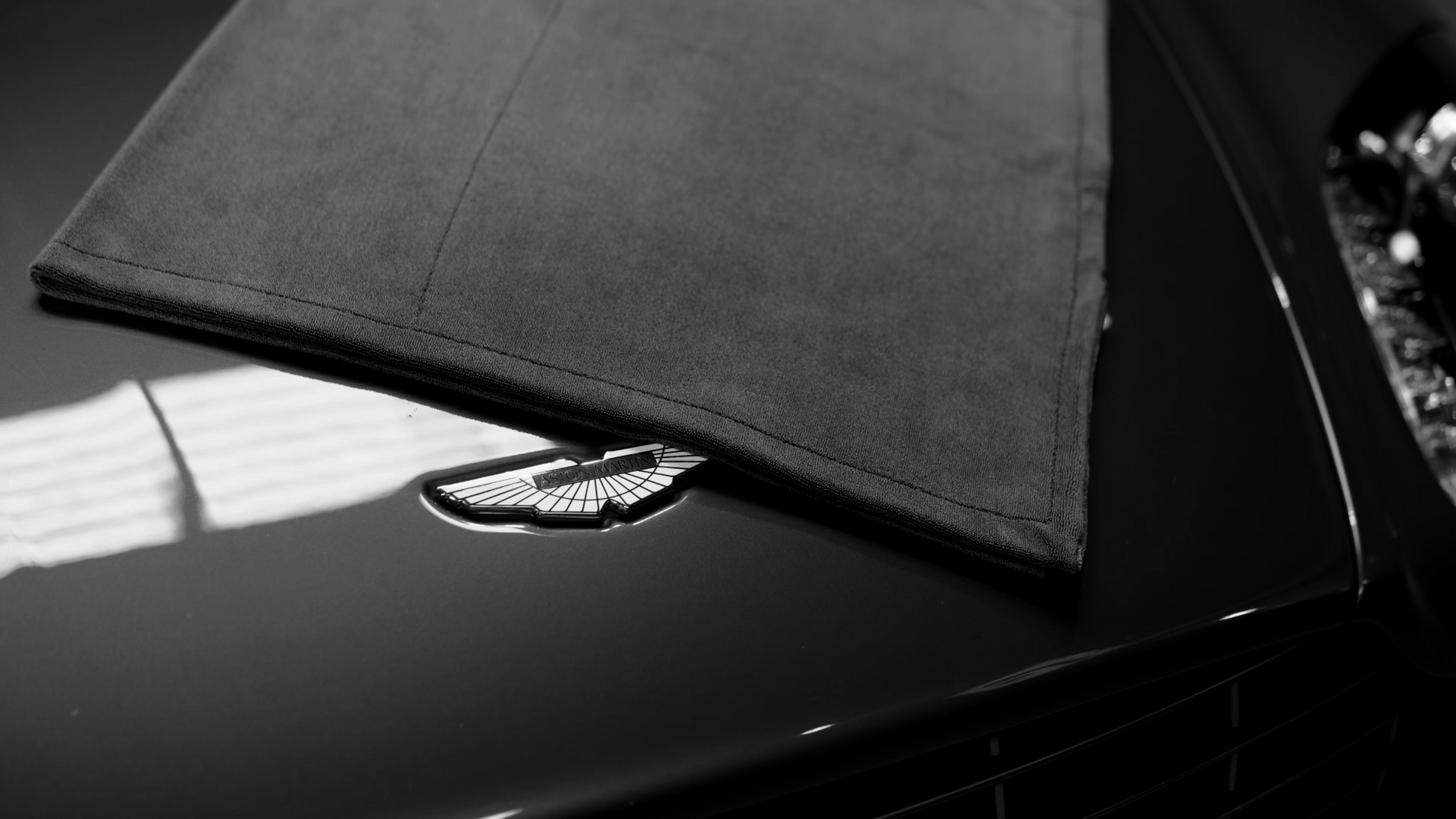 A close-up of an Aston Martin logo on the hood, partially covered by a FibreKing Gold Label 900 GSM Korean Drying Towel in black and white—perfect for showcasing luxury car care.