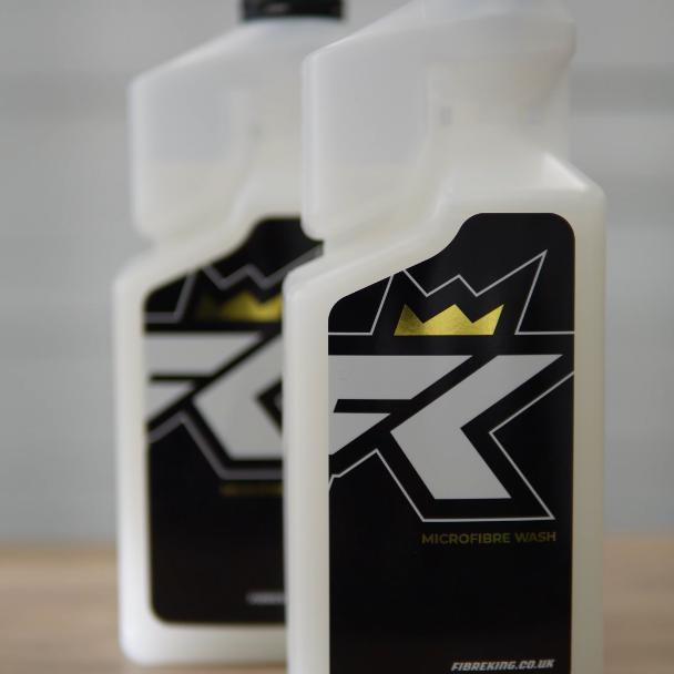 Two white FibreKing Microfibre Wash bottles with black labels and crown graphics sit on a wooden surface. This premium solution keeps your microfibre towels fresh and effective for detailing.
