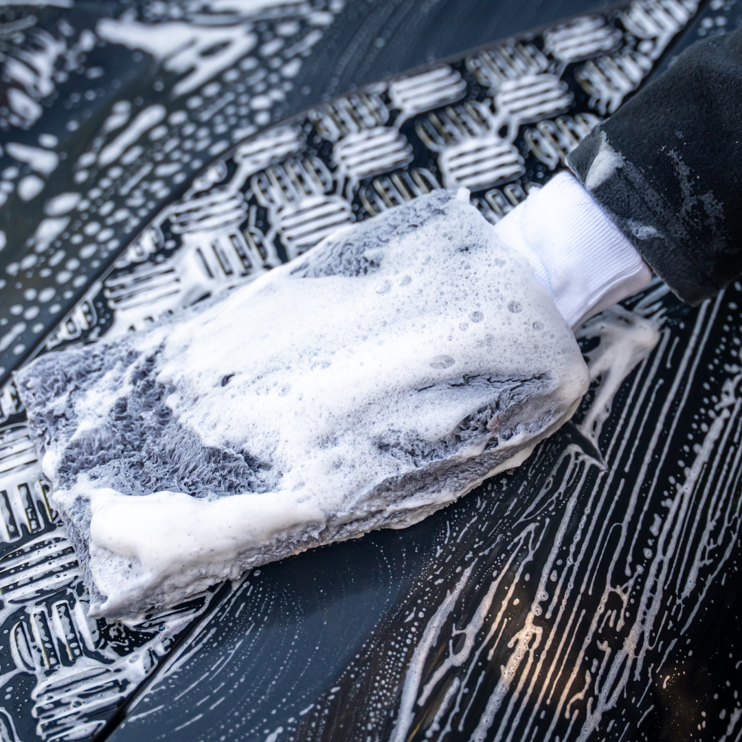 A hand in a black glove uses the FibreKing PS Korean Microfibre Mitt in grey to gently clean a textured black surface, creating visible suds and soap streaks for effective scratch prevention.