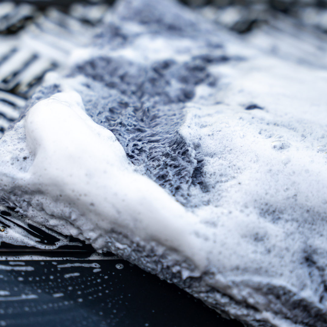 A close-up of the FibreKing PS Korean Microfibre Mitt in grey, covered in white foam on a dark wet surface with soap streaks, highlighting the ultra-soft fibres for optimal scratch prevention.