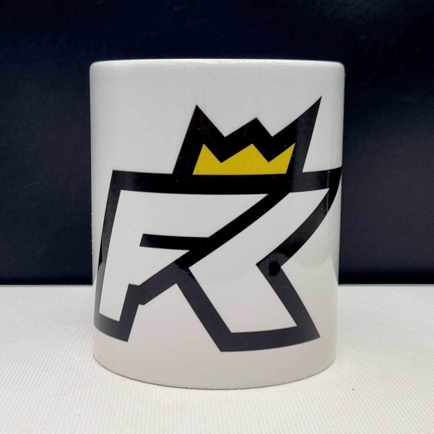 Enjoy your morning brew with the Fibreking Mug by FibreKing—a white handmade coffee mug featuring a bold black and yellow geometric RK logo topped with a crown, perfect for showing your team spirit.