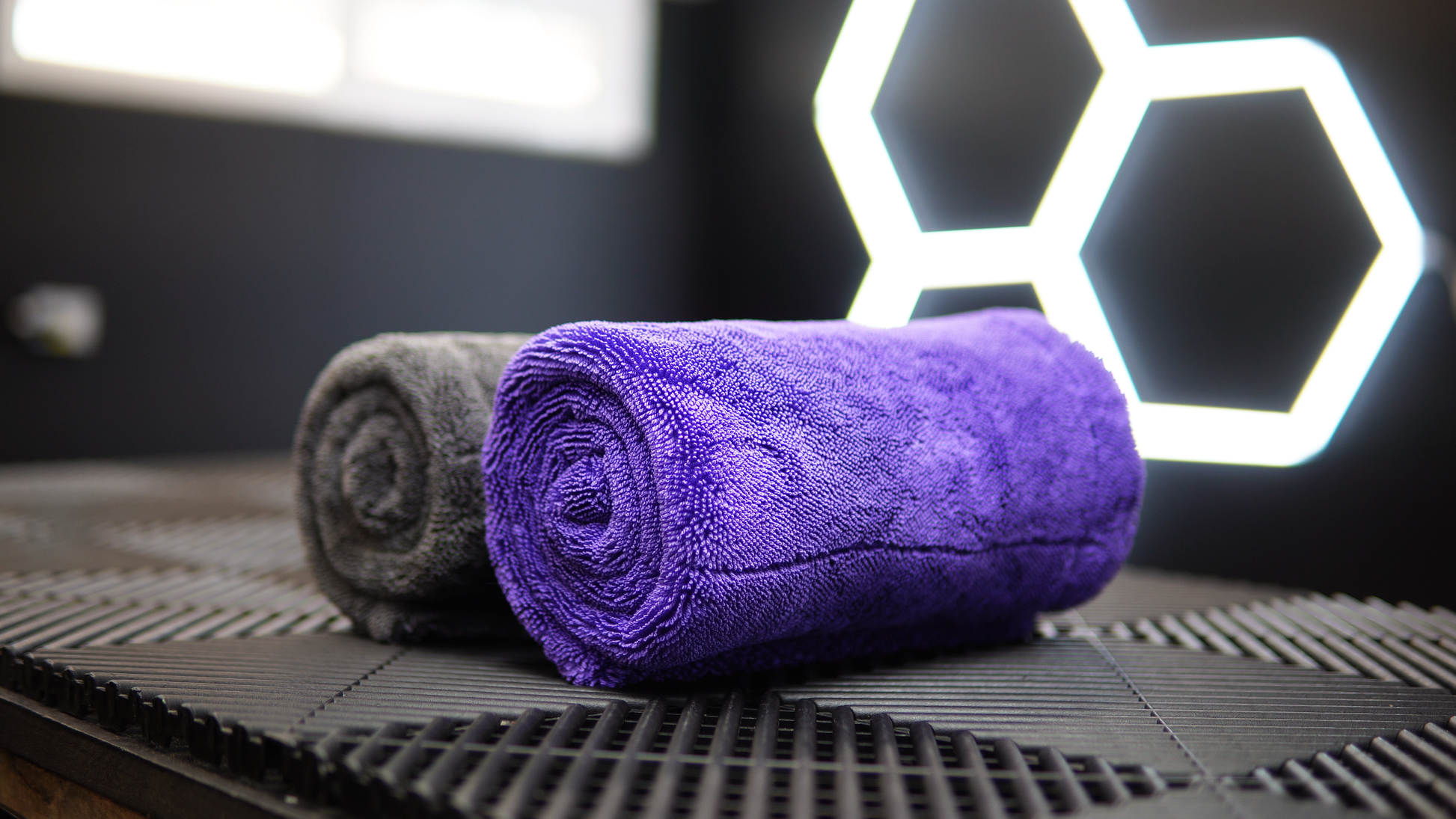 A rolled FibreKing Silver Label Drying Towel and a rolled purple car detailing towel sit side by side on a textured black surface with geometric patterns in the background.
