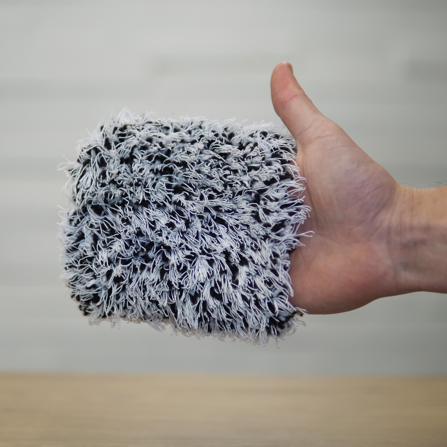 A hand holds the FibreKing Gold Label Korean Wheel Mitt—a gray and white microfiber mitt—over a wooden surface. The softly blurred background highlights its effective texture for gentle brake dust removal.