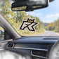 A FibreKing FK Air Freshener, featuring a black and white design with a crown and stylized letters, hangs from a car’s rearview mirror; the road and trees can be seen through the windshield.