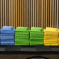 Stacks of blue, green, and yellow FibreKing Silver Label Microfibre Bundle cloths are neatly arranged on a black surface in front of wooden slats and geometric light fixtures.