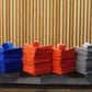 Stacks of neatly folded blue, orange, and gray FibreKing Gold Label Microfibre Bundle cloths, each topped with a rolled towel, are arranged on a black surface with a wooden slat background and geometric lighting.