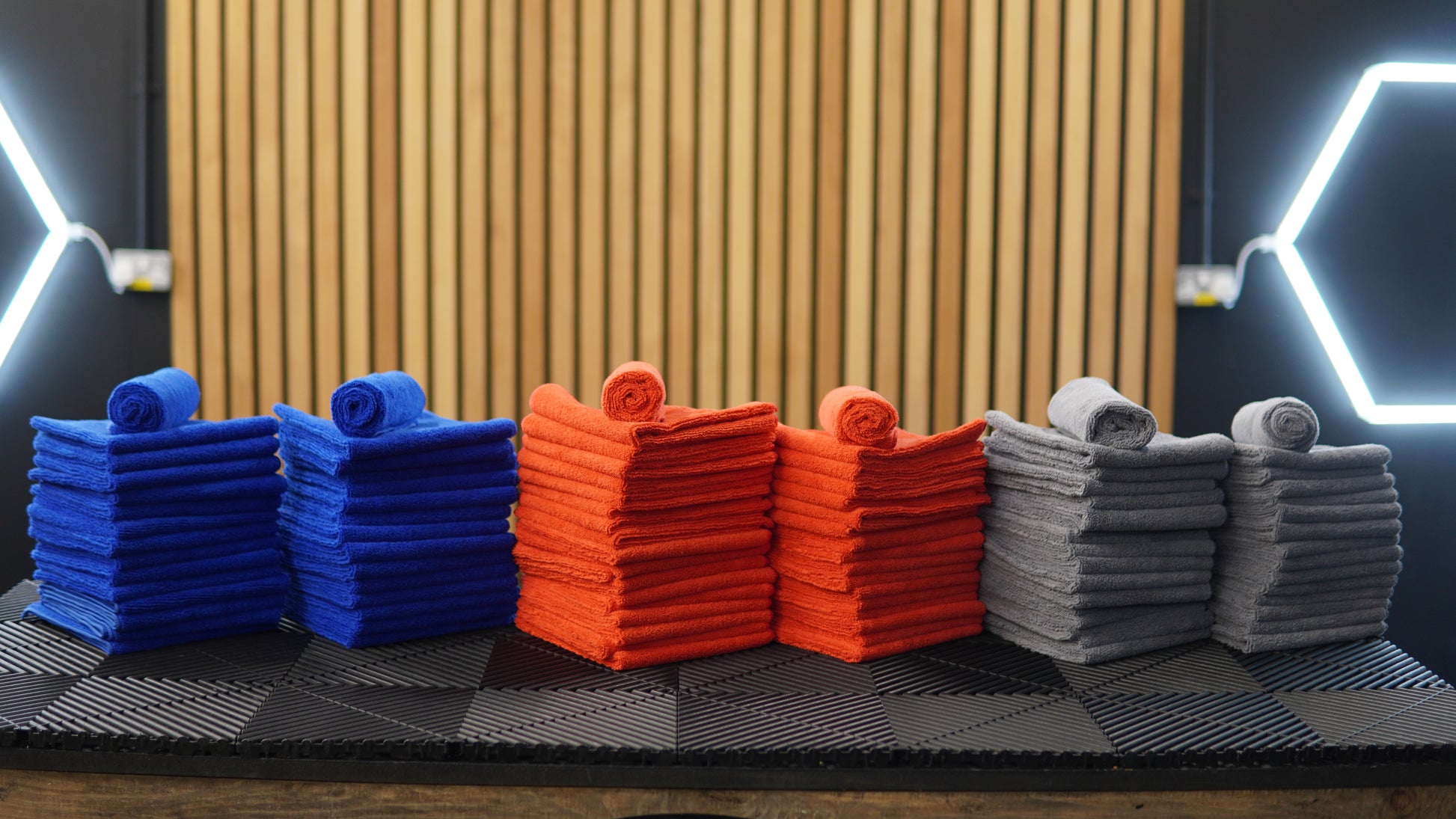 Stacks of neatly folded blue, orange, and gray FibreKing Gold Label Microfibre Bundle cloths, each topped with a rolled towel, are arranged on a black surface with a wooden slat background and geometric lighting.