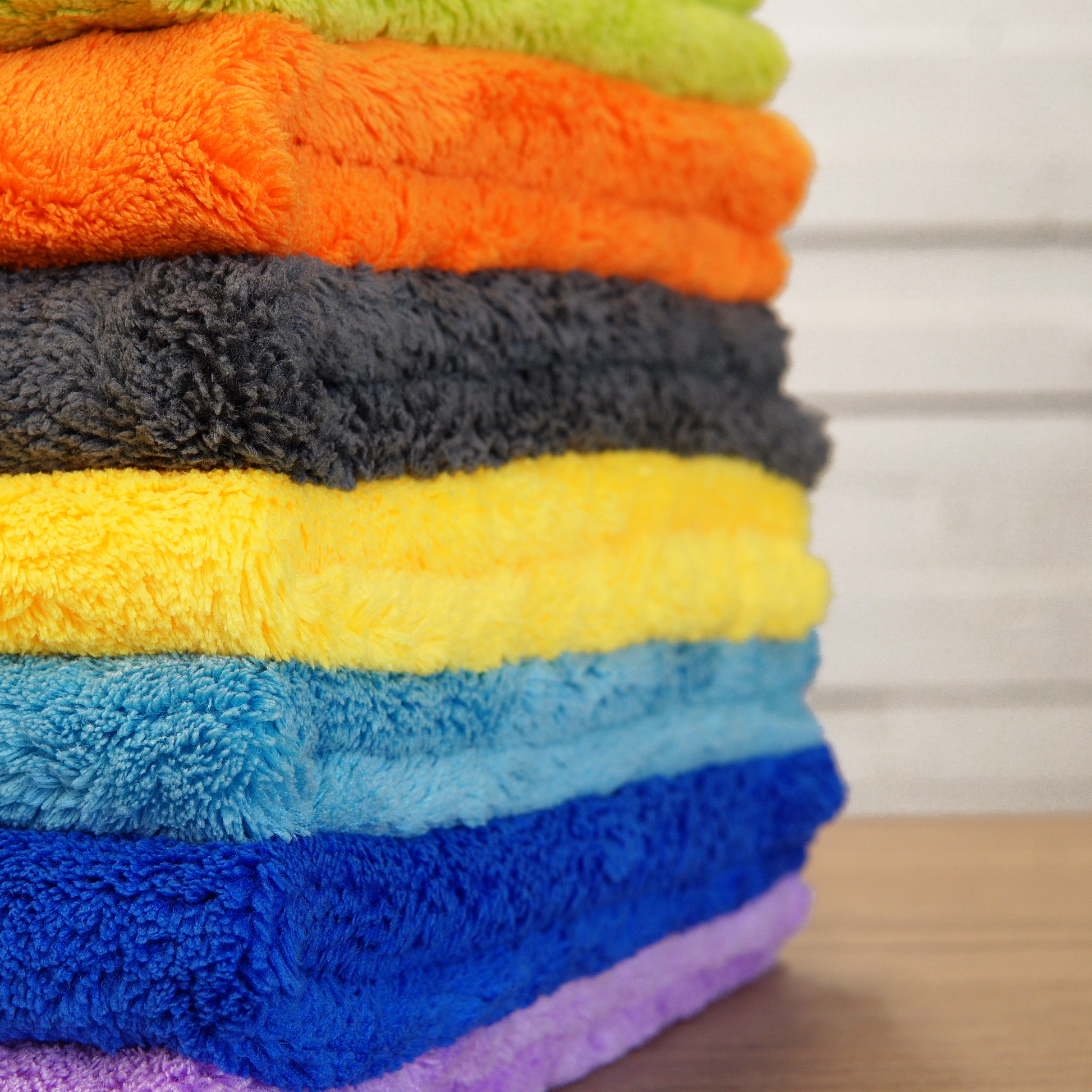 A stack of folded, colorful FibreKing Ultimate Detailer Cloth microfiber towels is displayed on a wooden surface against a softly blurred background.