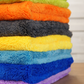 A stack of FibreKing Ultimate Detailer Cloth Multipack (3 or 5 pack) towels is neatly arranged in layers on a wooden surface, set against a light-striped background.