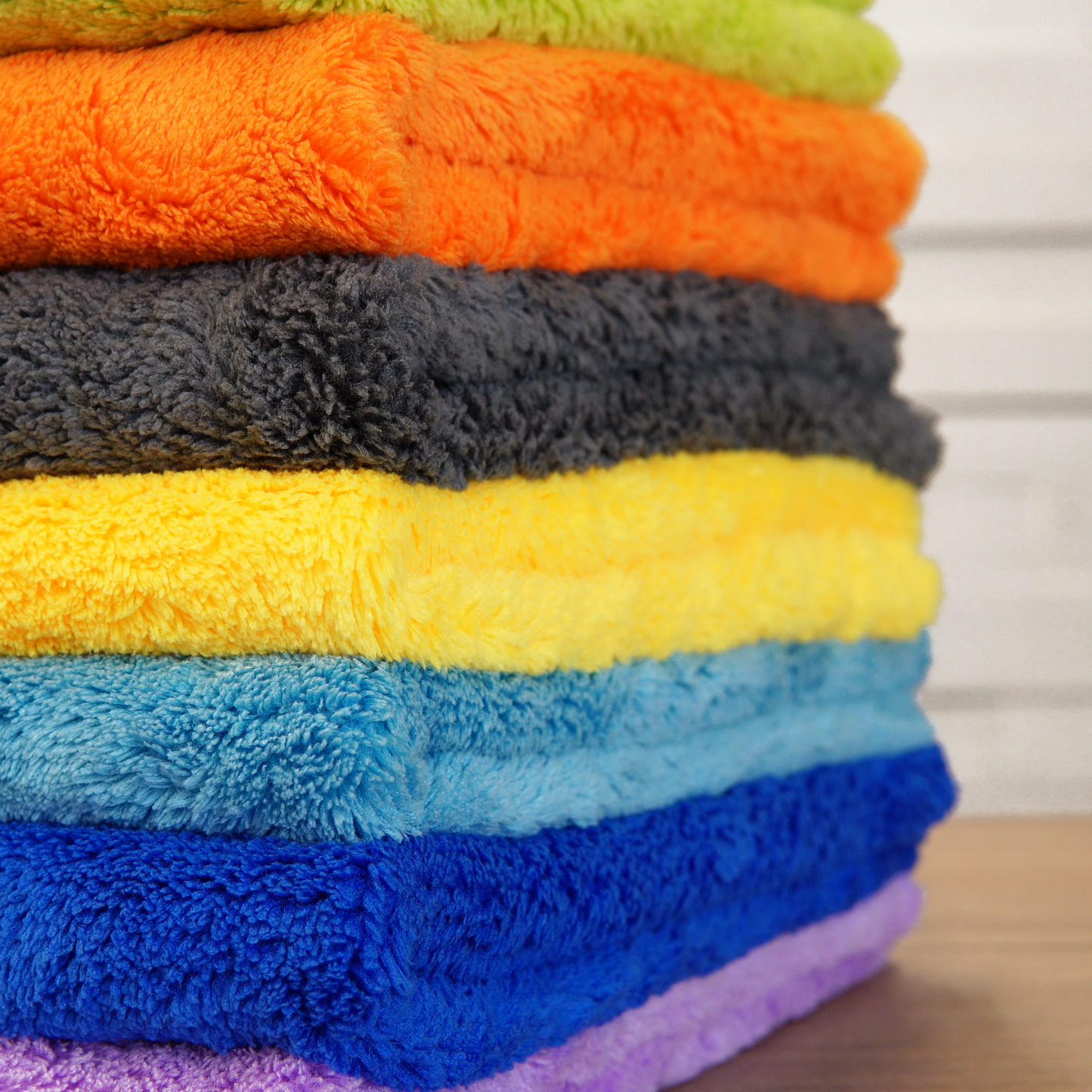 A stack of FibreKing Ultimate Detailer Cloth Multipack (3 or 5 pack) towels is neatly arranged in layers on a wooden surface, set against a light-striped background.