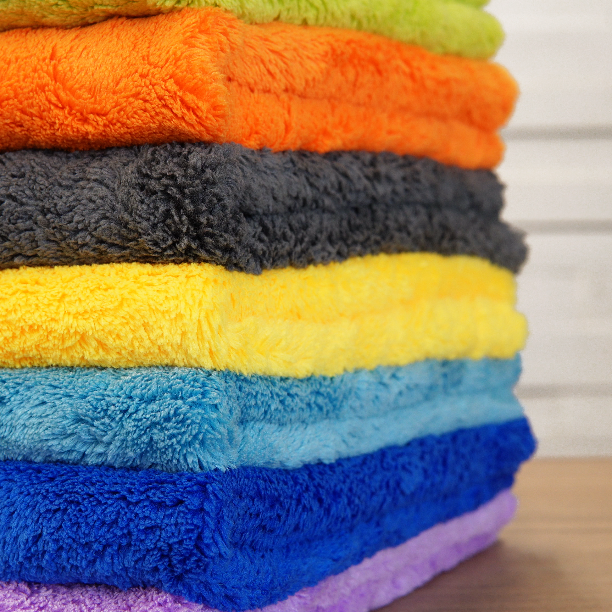 A stack of FibreKing Ultimate Detailer Cloth Multipack (3 or 5 pack) towels is neatly arranged in layers on a wooden surface, set against a light-striped background.