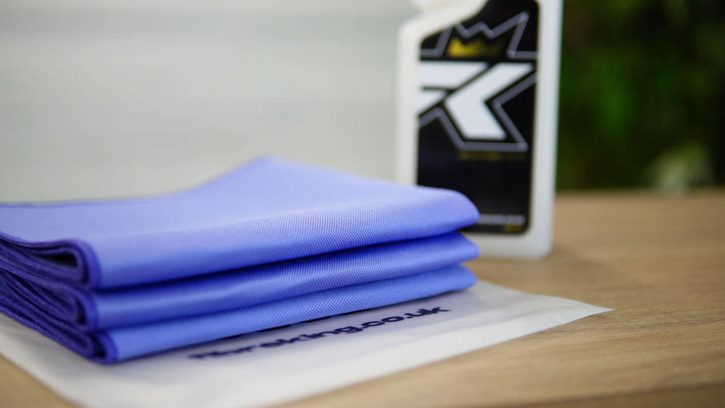 A neatly folded FibreKing Gold Label Korean Glass Cloth from the 3 Pack, known for high absorption and streak-free cleaning, sits on a plastic bag on a wooden surface, with part of a cleaning product bottle visible in the background.