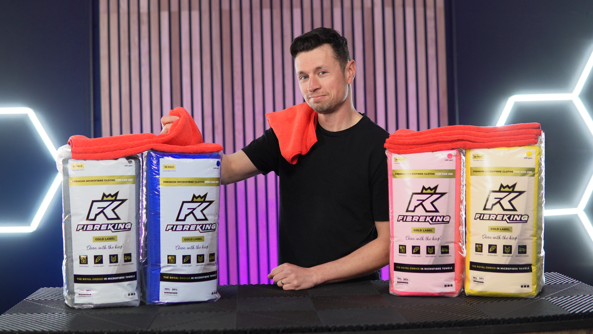 A man stands behind a table with four FibreKing Gold Label microfibre cloth packs, holding and draping an orange cloth from the FibreKing EPIC Bundle XL on his shoulder and arm.