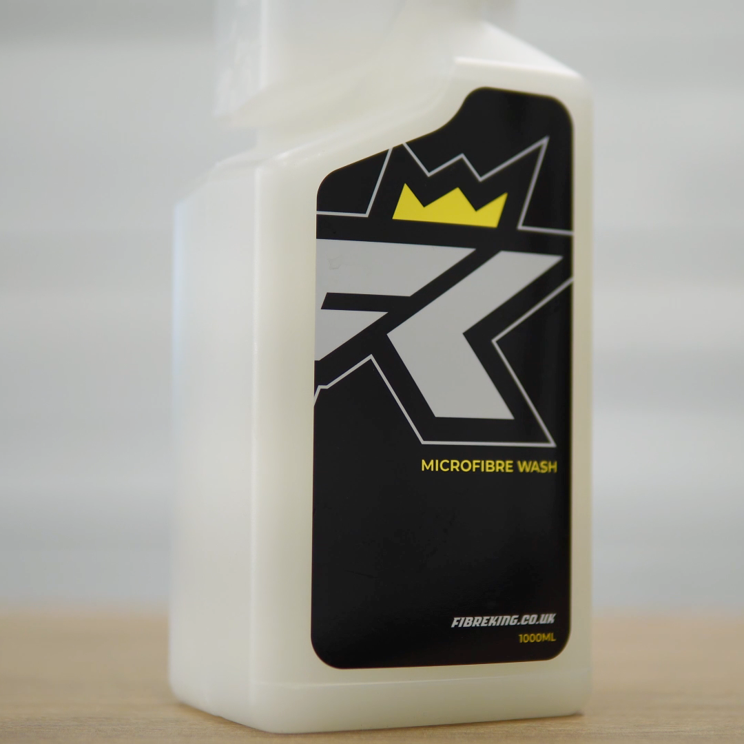 A plastic FibreKing Microfibre Wash bottle, designed for caring for microfibre towels, features a black, white, and yellow design and sits on a wooden surface against a light background.