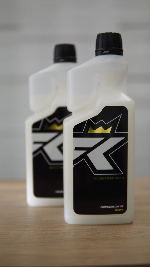 Two white FibreKing Microfibre Wash bottles with black labels and crown graphics sit on a wooden surface. This premium solution keeps your microfibre towels fresh and effective for detailing.