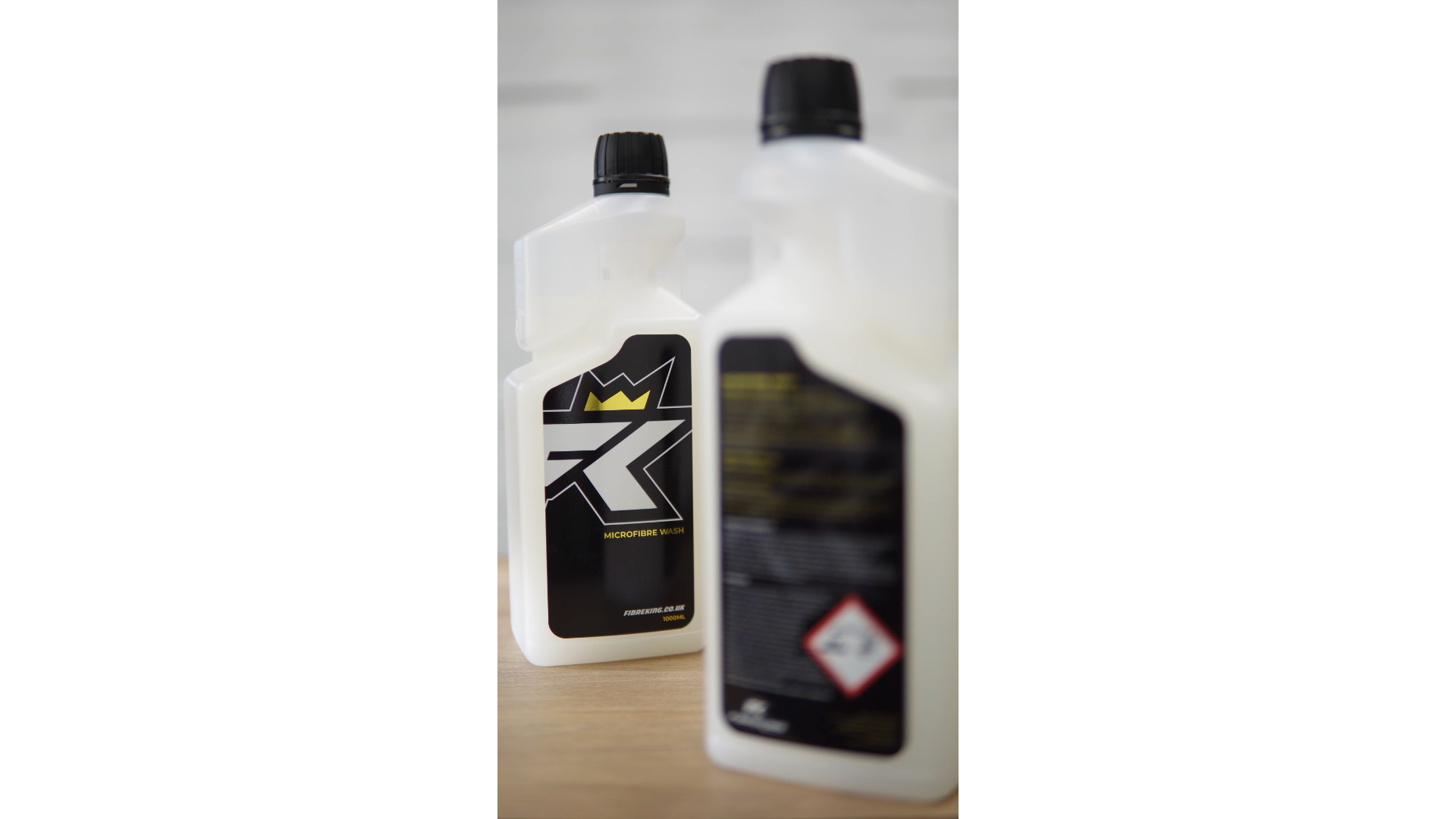 Two white FibreKing Microfibre Wash bottles with black caps—one focused showing the stylized R logo and crown, the other blurred with hazard symbols—ideal for caring for microfibre towels or as a detailing towel cleaner.