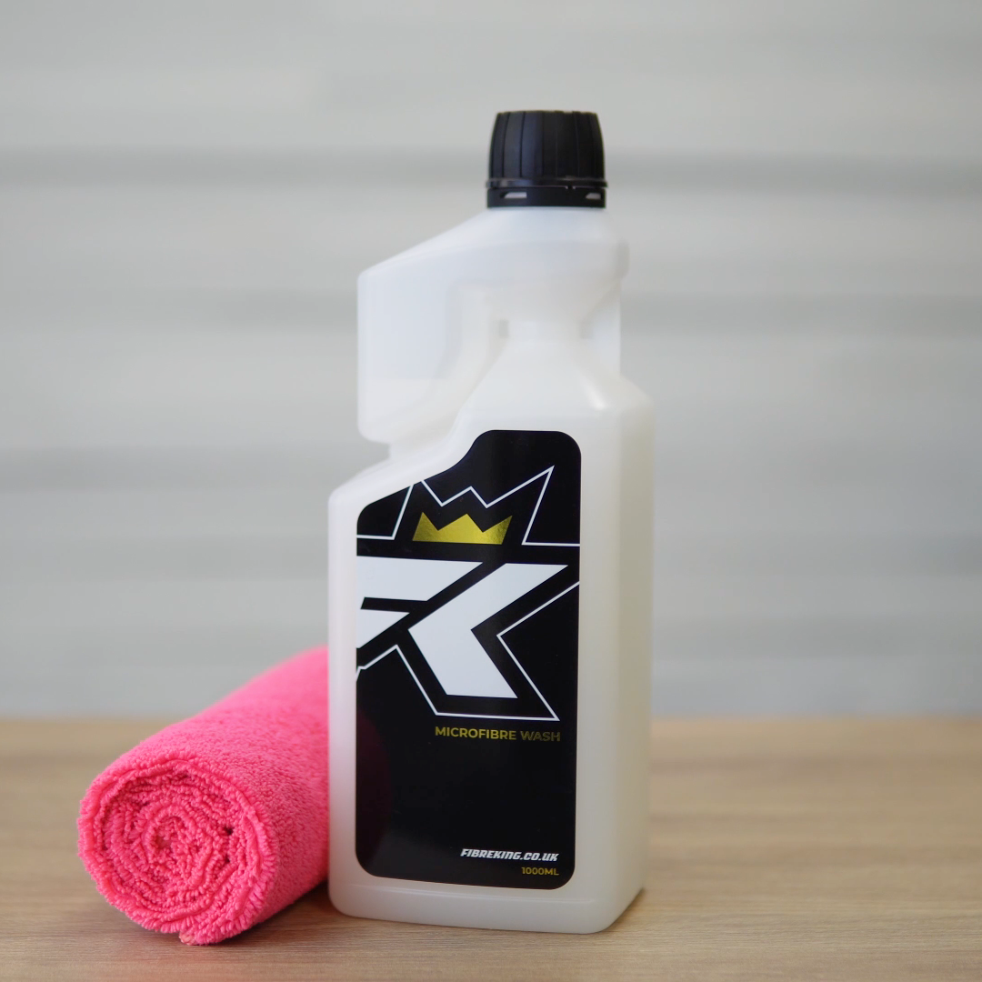 A bottle of FibreKing Microfibre Wash with a black label and gold crown logo stands next to a rolled pink microfibre cloth on a wooden surface, ideal for caring for your microfibre towels.