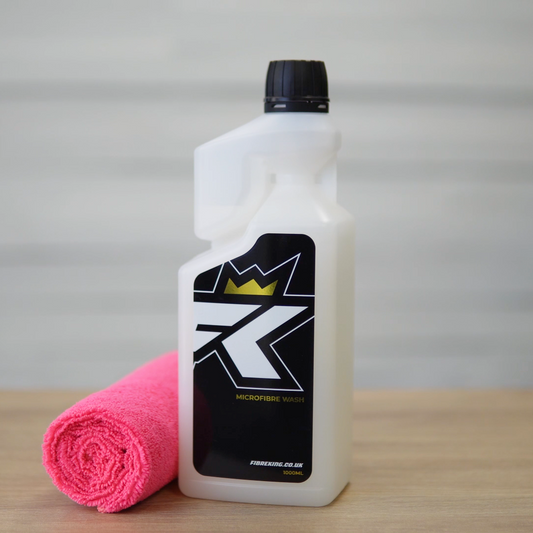 A bottle of FibreKing Microfibre Wash with a black label and gold crown logo stands next to a rolled pink microfibre cloth on a wooden surface, ideal for caring for your microfibre towels.
