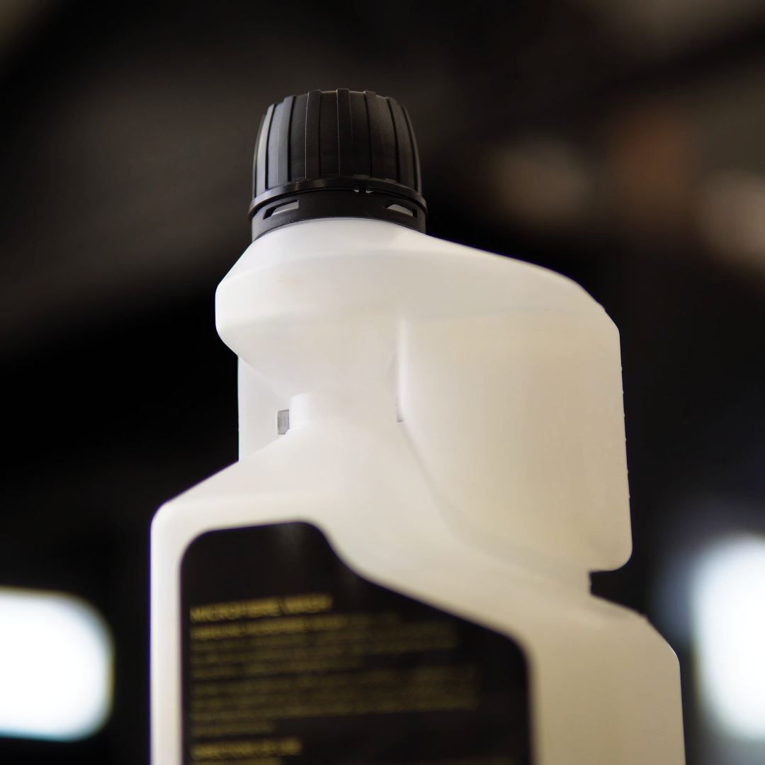 A close-up of FibreKing Microfibre Wash, a white plastic bottle with a black cap and black label, ideal for microfibre towel care, shown against a blurred background.