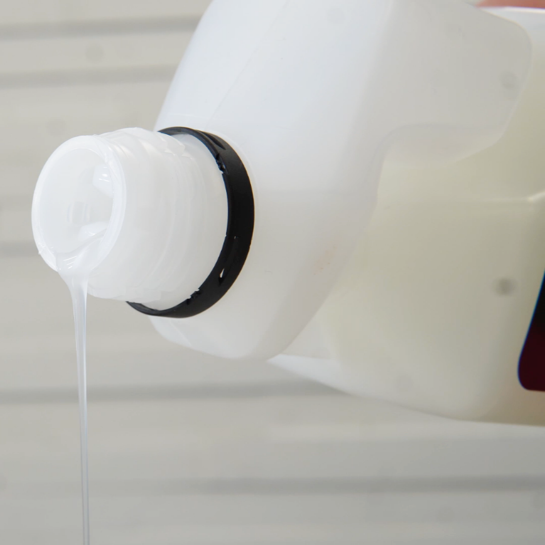 A close-up of FibreKing Microfibre Wash, a white plastic bottle pouring clear liquid, perfect for microfibre towel care, with a blurred light background.