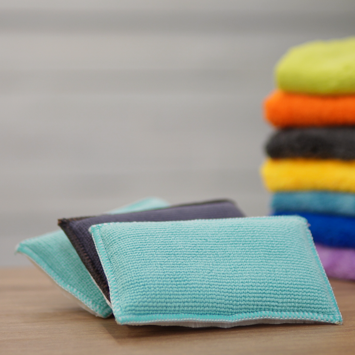 Two rectangular mint blue Scrub Pads by FibreKing rest on a wooden surface, with colorful folded towels in the background—an ideal addition to any car cleaning or detailing kit.