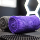 A rolled FibreKing Silver Label Drying Towel and a rolled purple car detailing towel sit side by side on a textured black surface with geometric patterns in the background.