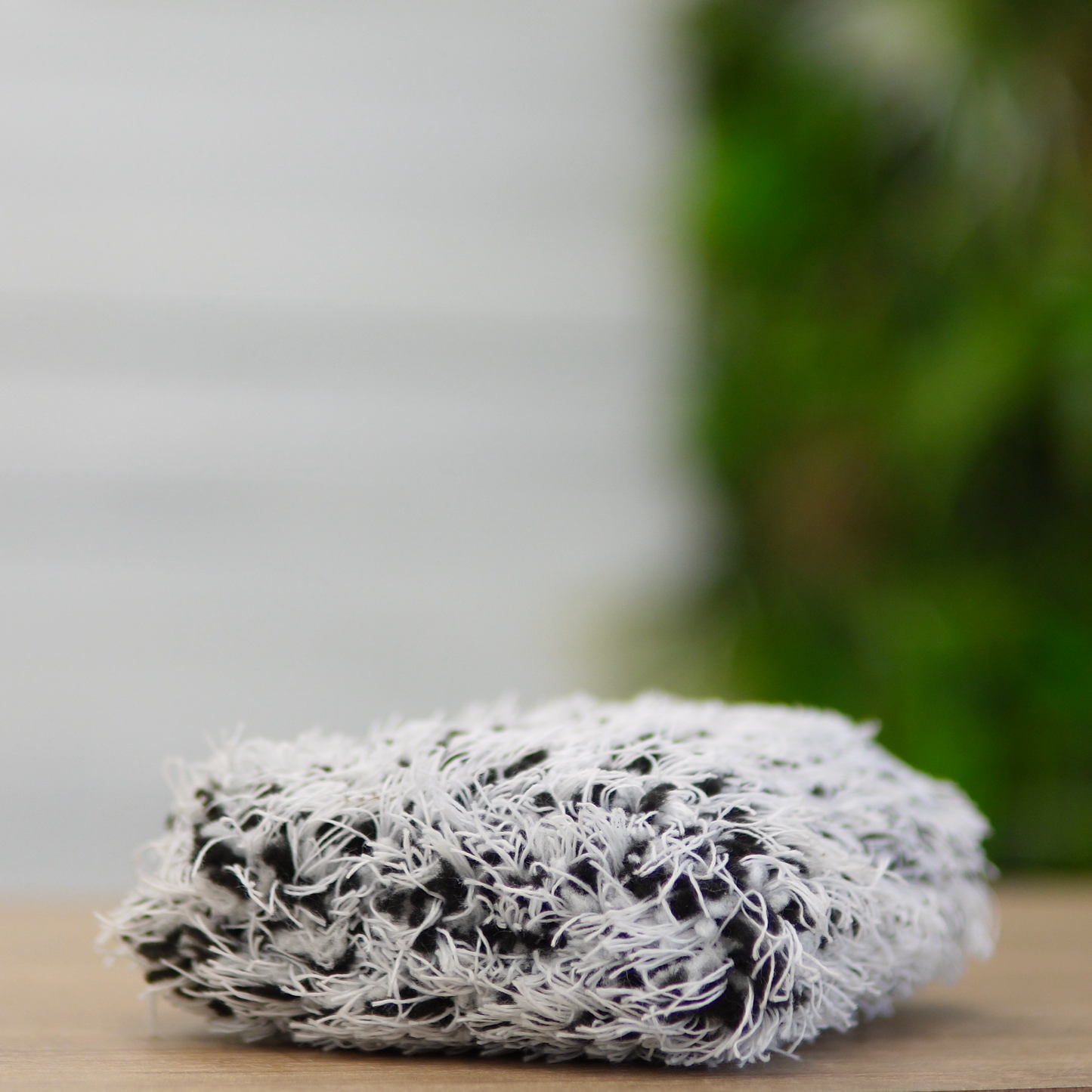 A close-up of the FibreKing Gold Label Korean Wheel Mitt—a gray, rectangular cleaning mitt with frayed fibers for brake dust removal—rests on a wooden surface, set against a blurred green background.