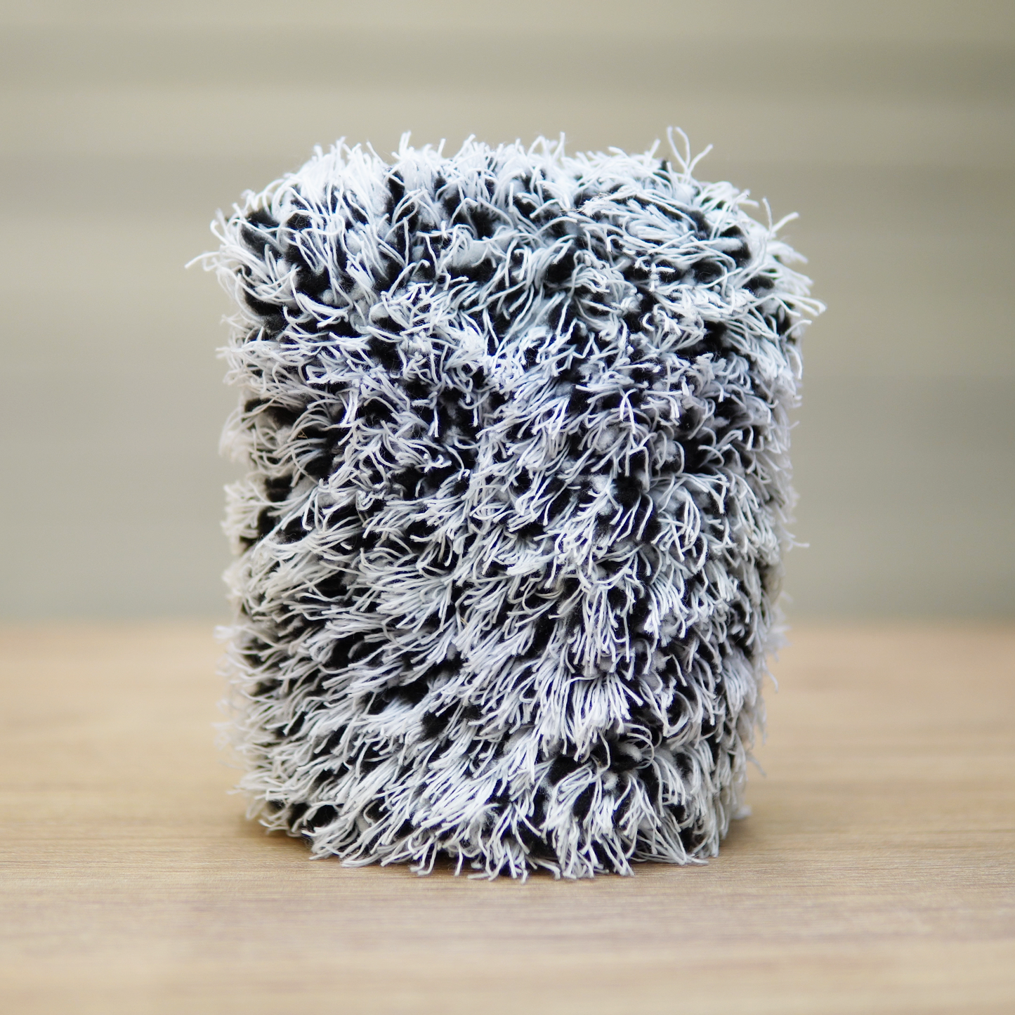 The FibreKing Gold Label Korean Wheel Mitt, a gray cylindrical chenille microfiber mitt, stands upright on wood with a blurred background—ideal for effective brake dust removal.