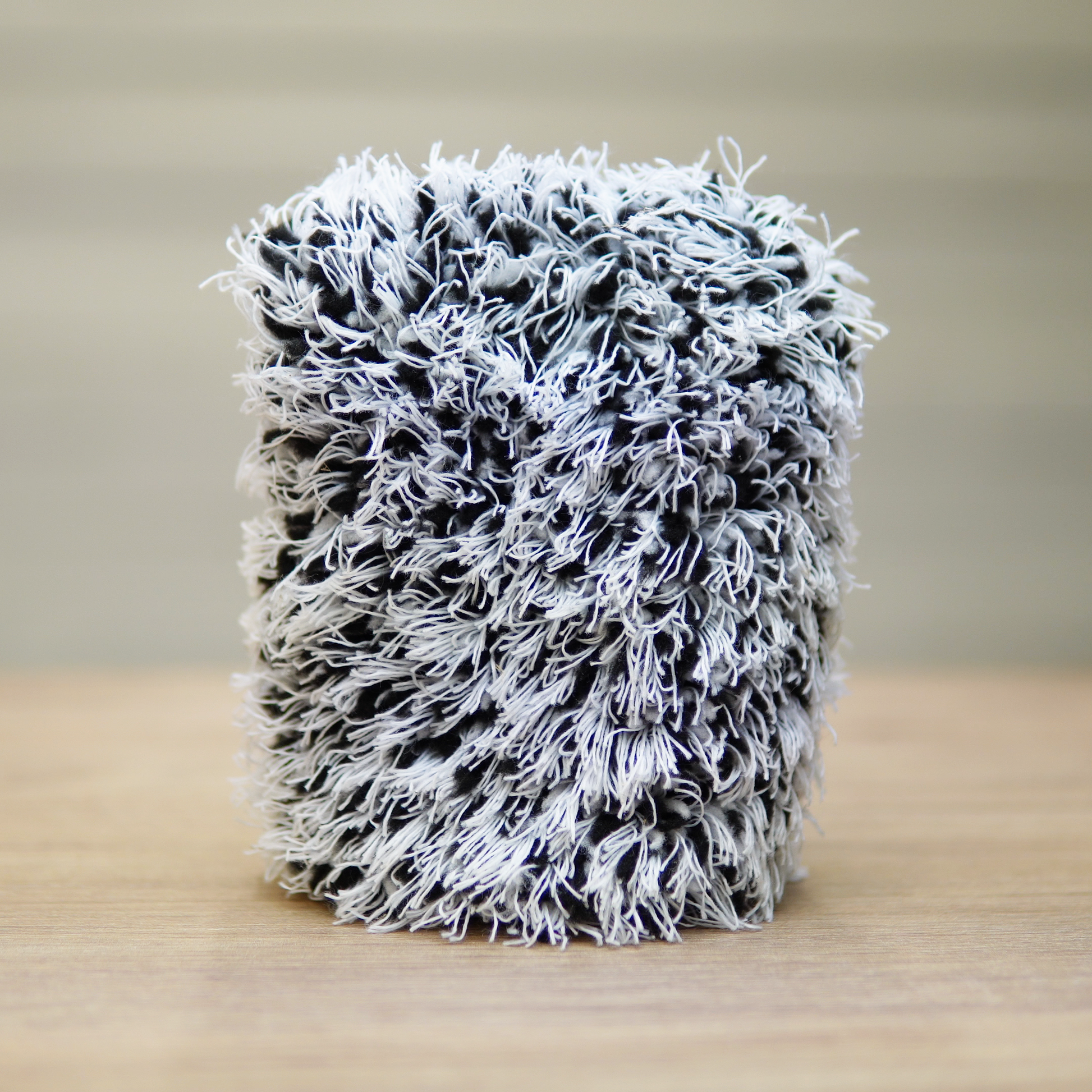 The FibreKing Gold Label Korean Wheel Mitt, a gray cylindrical chenille microfiber mitt, stands upright on wood with a blurred background—ideal for effective brake dust removal.