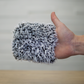 A hand holds the FibreKing Gold Label Korean Wheel Mitt—a gray and white microfiber mitt—over a wooden surface. The softly blurred background highlights its effective texture for gentle brake dust removal.